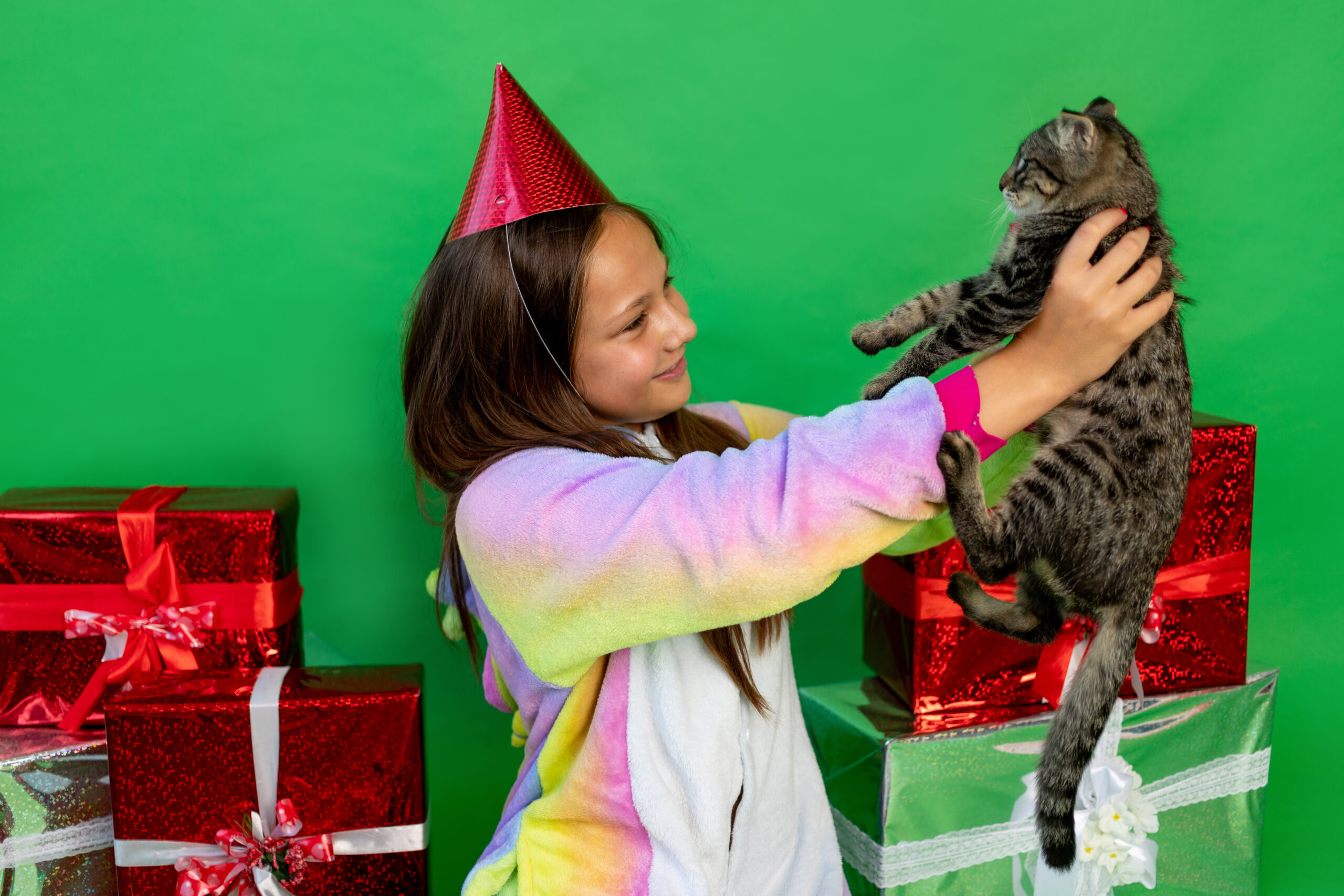 Uma Menina que gosta de gatos segurando um gato e rodeada de Presentes presentes para que gosta de gatos. No fundo, uma parede verde, a menina está sorrindo e olhando para o gato.